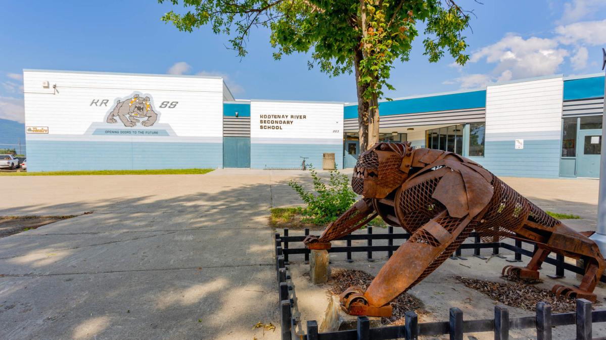 A flat-roofed blue and white school building against a blue sky with a green, leafy tree and a rusty metal sculpture of a bulldog in the foreground.