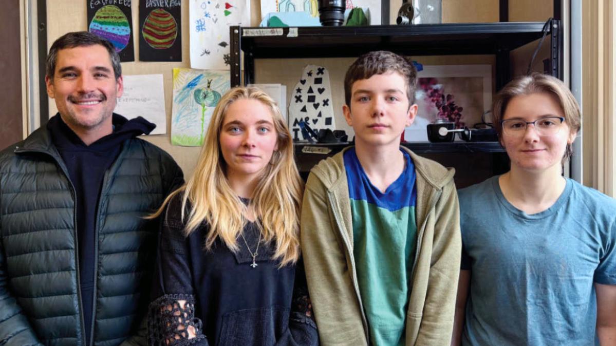 A man in a black down sweater stands beside three teens dressed in casual clothing in a school office.