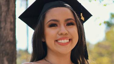 Young woman with black hair and big smile wearing graduation cap and light pink strappy dressed seated in outdoor setting in sunshine looking directly at viewer.