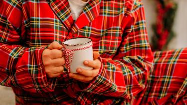Person in red plaid pyjamas holding a white mug with a red and while striped string tied around it.