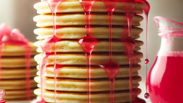 A tall stack of pancakes on a medium stoneware plate with pink syrup flowing down the sides and a jug of pink syrup in the background. 