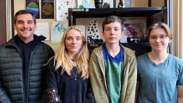 A man in a black down sweater stands beside three teens dressed in casual clothing in a school office.